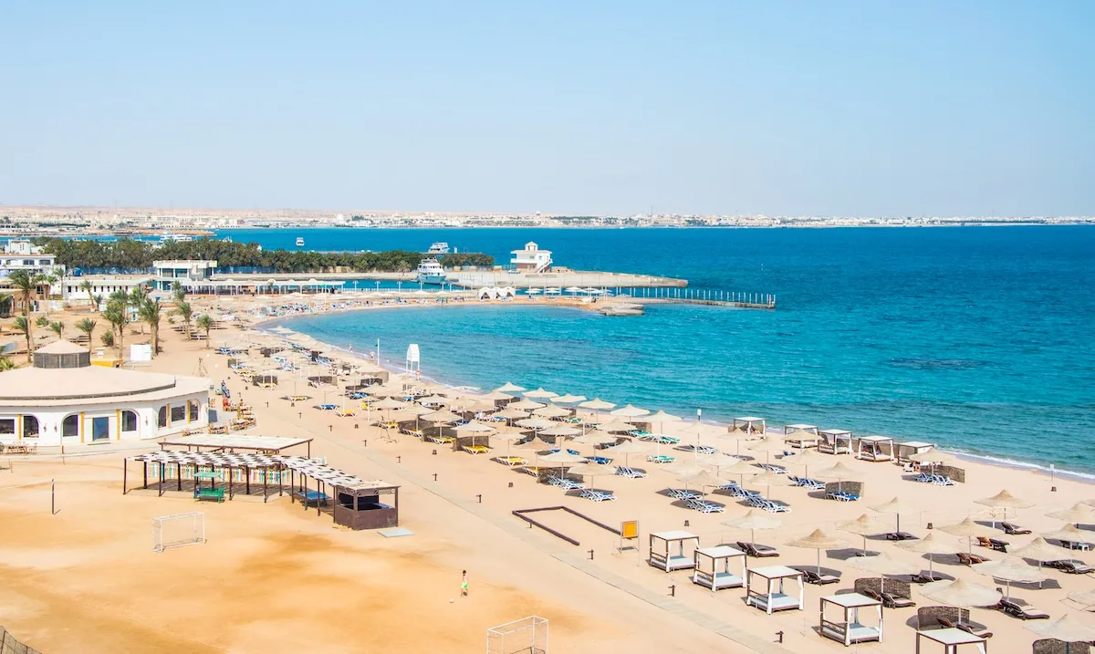 A hotel beach in Sahl Hasheesh with straw parasols, sunbeds and white cabanas along a turquoise bay, and a pier in the distance